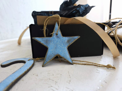 Box 2: A handcrafted blue star-shaped ceramic ornament with twine is displayed before a luxury gift box with tan ribbon and black tissue paper—perfect for Christmas gifts on a white surface.