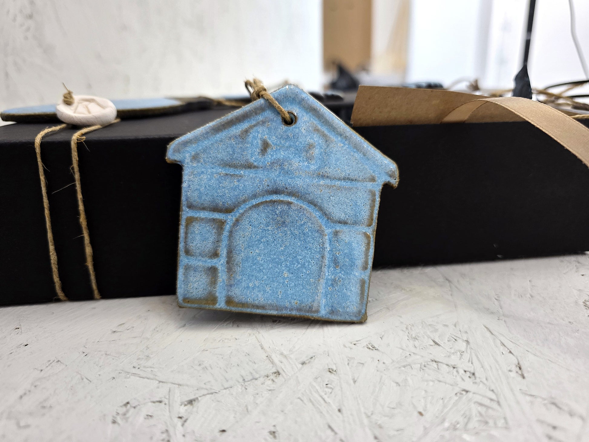 The "Box 3" handcrafted ceramic house ornament with a blue glaze hangs on twine in front of a luxury gift box tied with string, set against a white textured background with blurred objects.