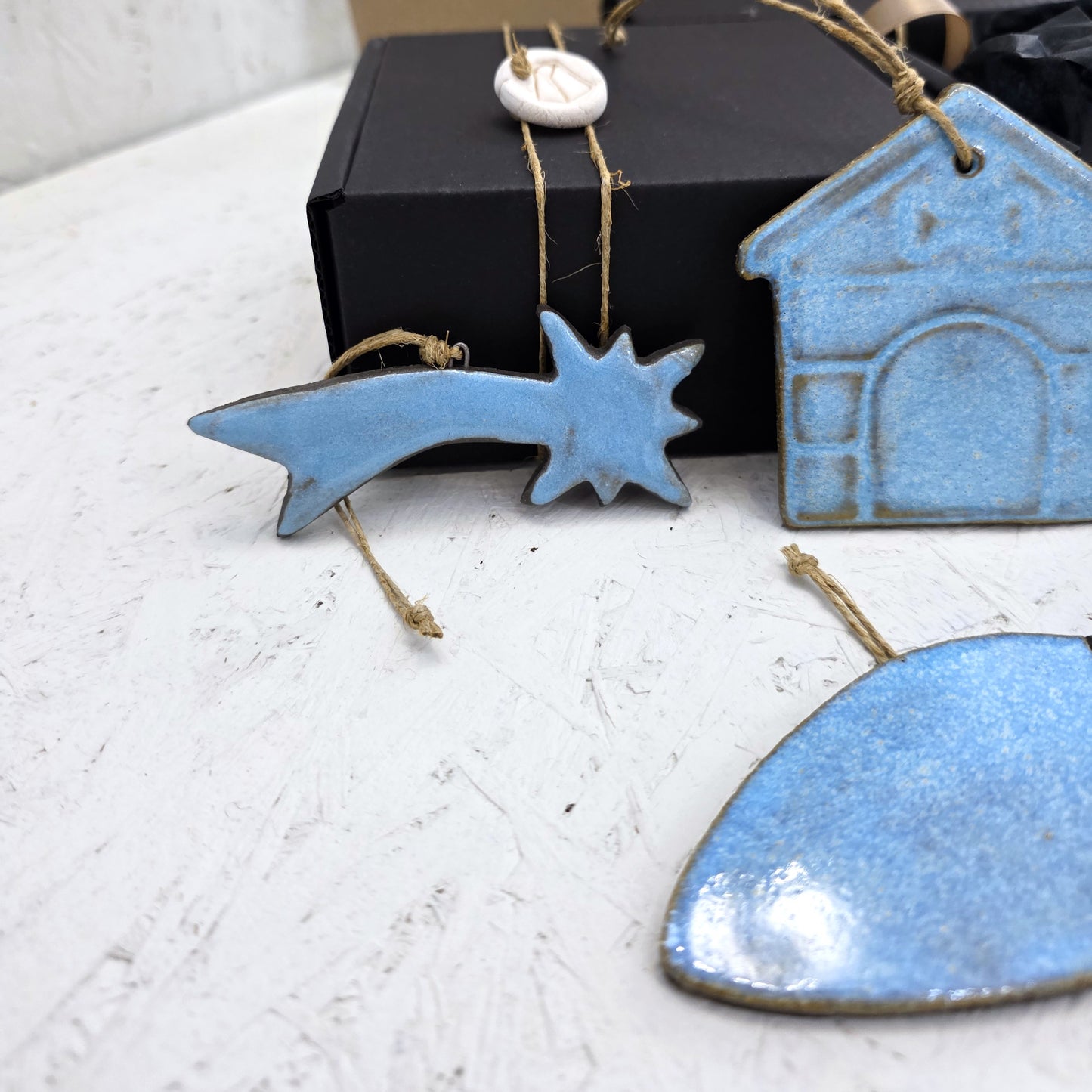 A close-up of Box 3: handcrafted light blue ceramic comet, house, and heart ornaments with twine loops on a white surface. Ideal Christmas gifts, presented in a luxury gift box partially visible in the background.