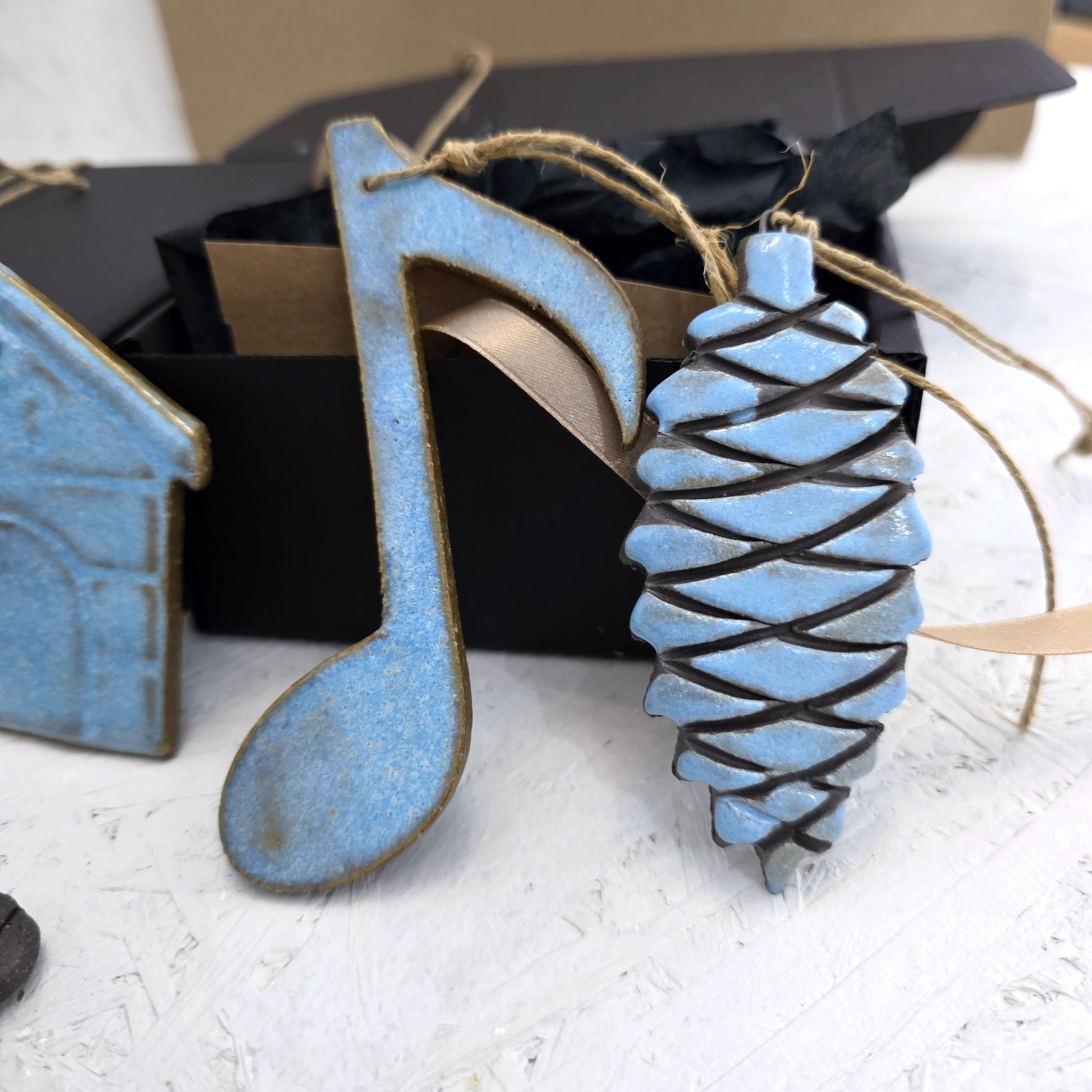 Box 3 features a handcrafted blue ceramic music note and pinecone ornament, both with brown details and twine for hanging, displayed near a luxury gift box—an ideal Christmas gift.