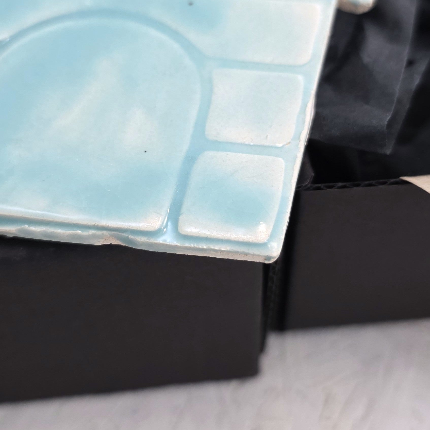A close-up of Box 4, a light blue handcrafted ceramic ornament tile with an embossed abstract pattern, displayed on the edge of a luxury gift box lined with black tissue paper atop a white, slightly textured surface.