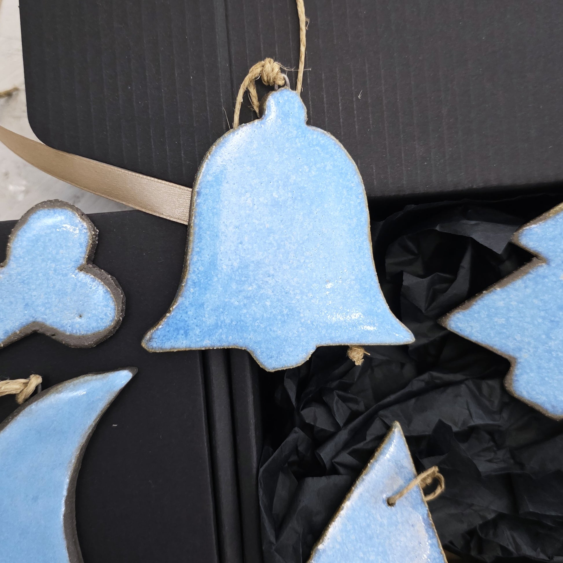 A close-up of Box 5, featuring blue handcrafted ceramic bell-shaped ornaments with twine loops, set on black crinkled paper in a luxury gift box—an ideal choice for Christmas gifting.