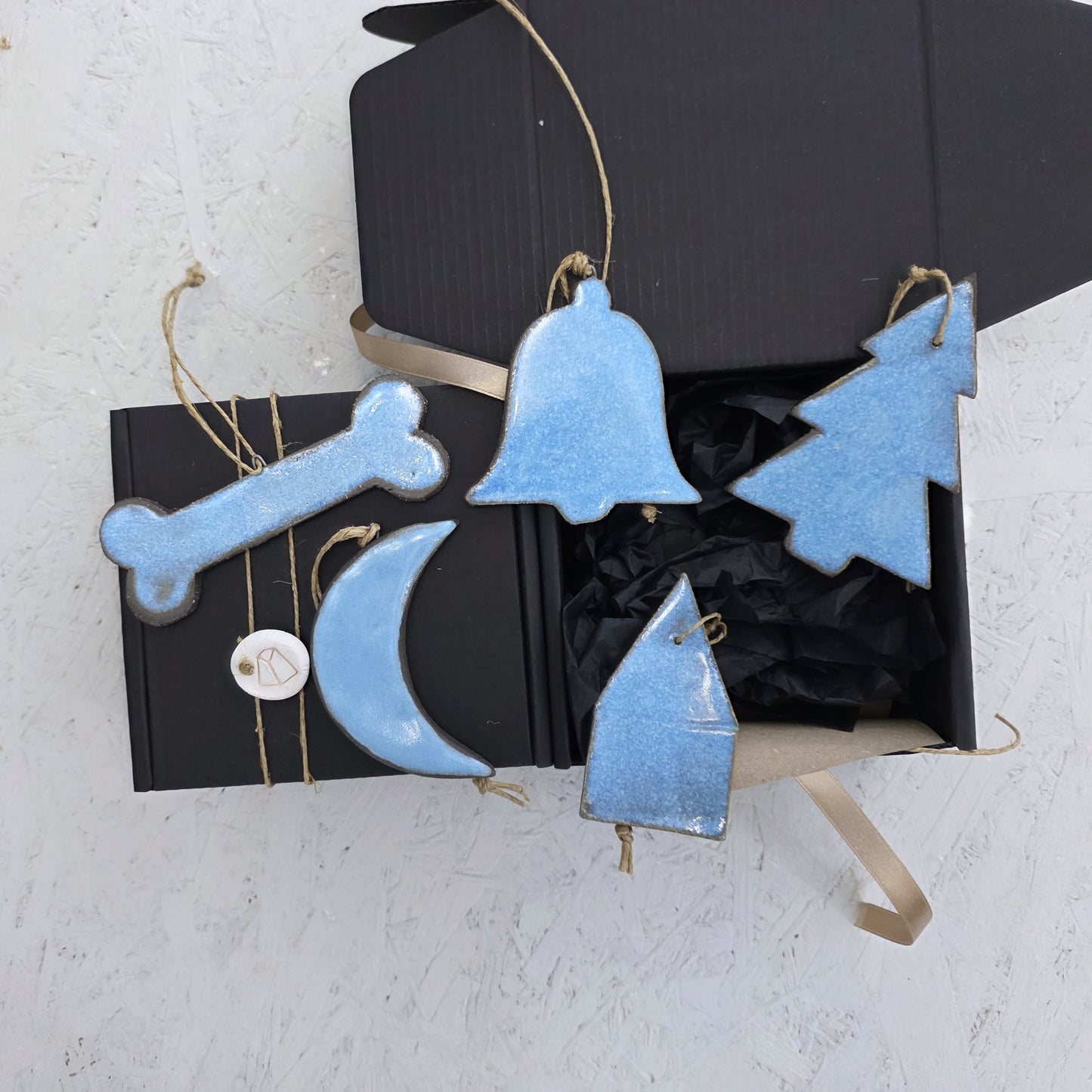 Box 5 includes four blue handcrafted ceramic ornaments—a bone, tree, moon, and bell—each hanging from twine loops and displayed with a luxury ribboned gift box. Ideal for Christmas gifting on a light textured surface.
