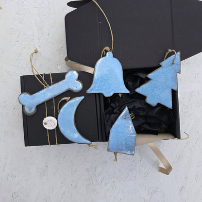 Box 5 includes four blue handcrafted ceramic ornaments—a bone, tree, moon, and bell—each hanging from twine loops and displayed with a luxury ribboned gift box. Ideal for Christmas gifting on a light textured surface.