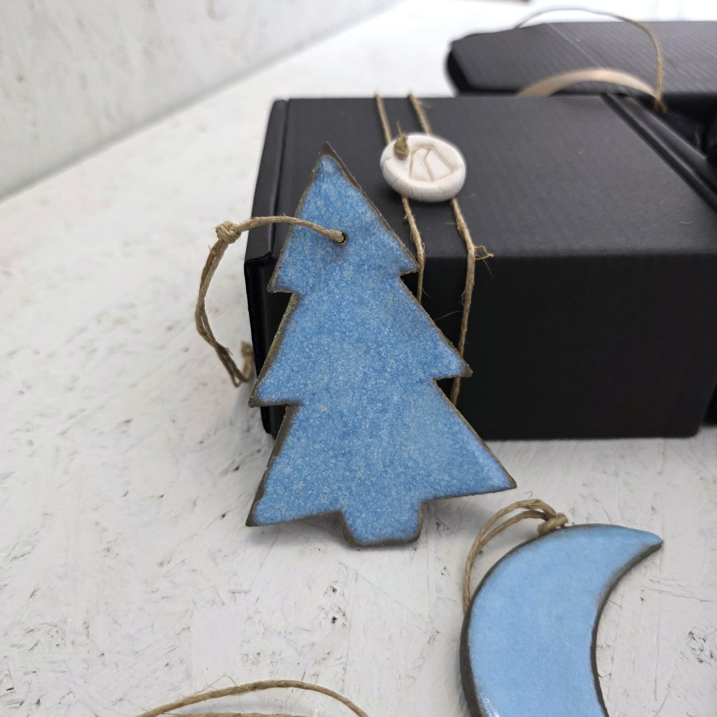 A handcrafted blue ceramic ornament with twine from Box 5 rests on a white surface beside a luxury gift box. A crescent moon ornament and a star-tag add charm, making it perfect for Christmas gifting.