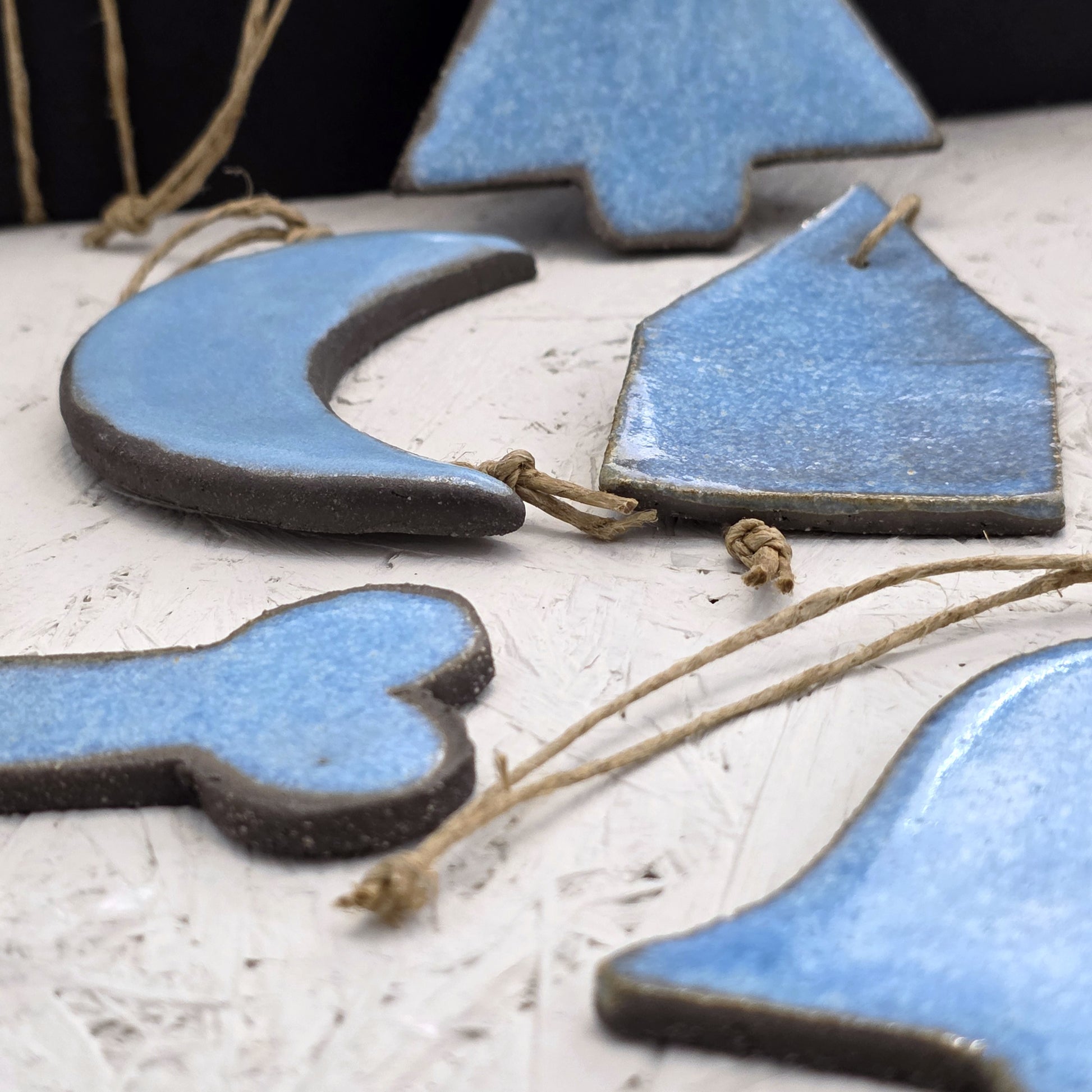 Box 5 features handcrafted ceramic ornaments shaped like a crescent moon, house, bell, and cross. Glazed in light blue and tied with twine, they’re perfect for Christmas gifting and displayed on a textured white surface.