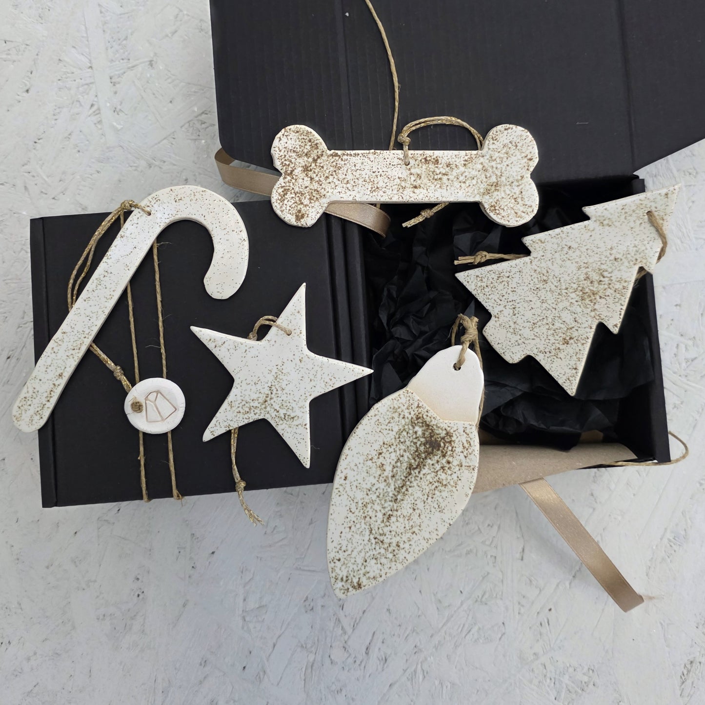 Box 7 is a black handcrafted gift box with luxury ceramic ornaments—speckled white and handmade—in candy cane, star, dog bone, tree, and light bulb shapes, ideal for Christmas gifting and displayed on a textured white surface.