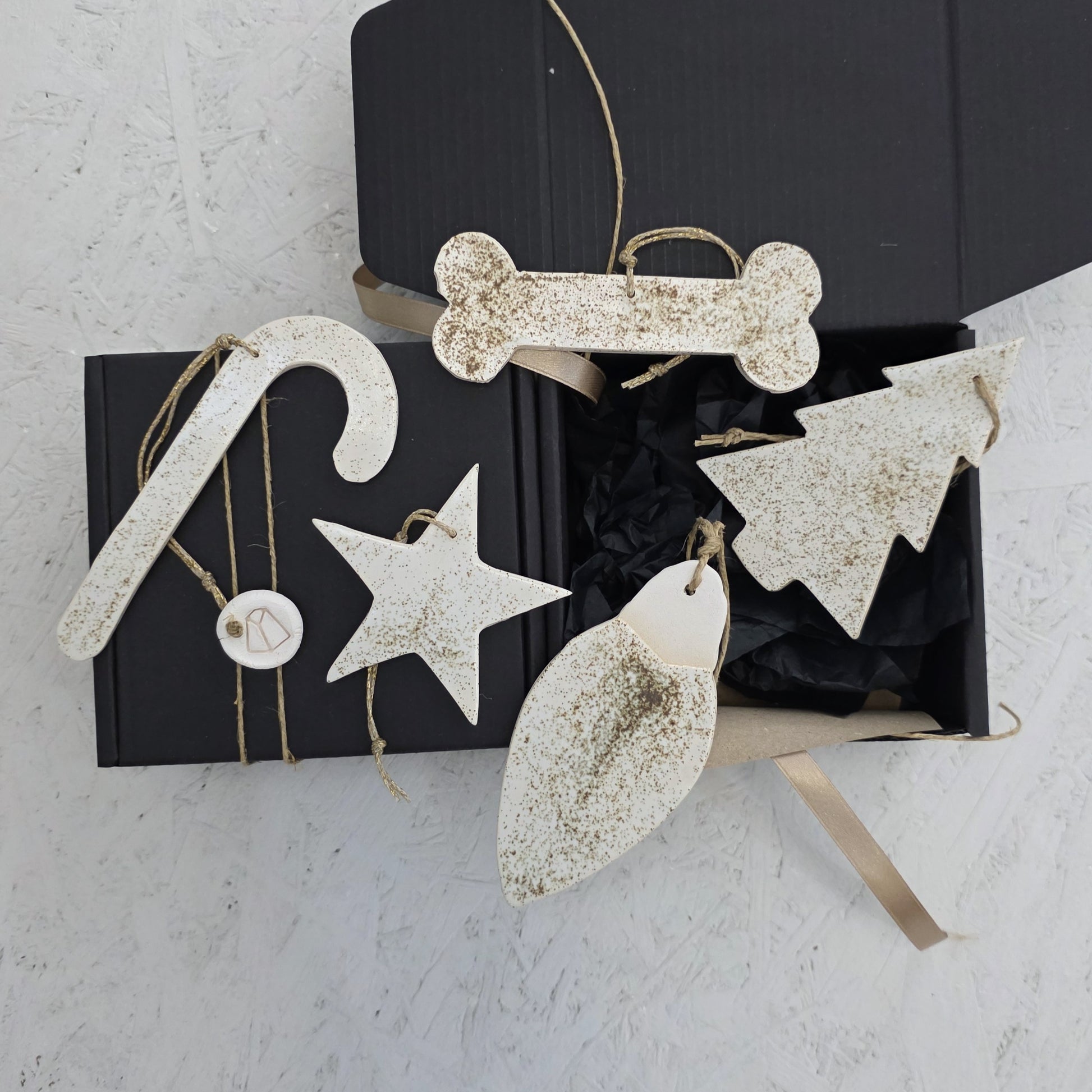 Box 7 is a black handcrafted gift box with festive white luxury ceramic ornaments—candy cane, star, tree, bone, and leaf—sprinkled with gold glitter and tied with twine. The box sits partially open on a light textured surface.