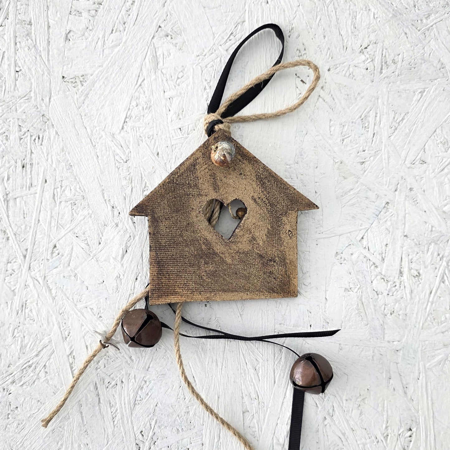 The Lucky charm House is a rustic brown wooden ornament with a heart-shaped cutout, twine and black ribbon, accented by two metal bells, inspired by festive traditions and displayed on a textured white surface.