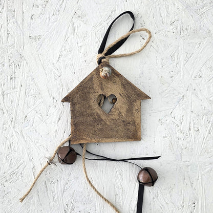 The Lucky charm House is a rustic brown wooden ornament with a heart-shaped cutout, twine and black ribbon, accented by two metal bells, inspired by festive traditions and displayed on a textured white surface.