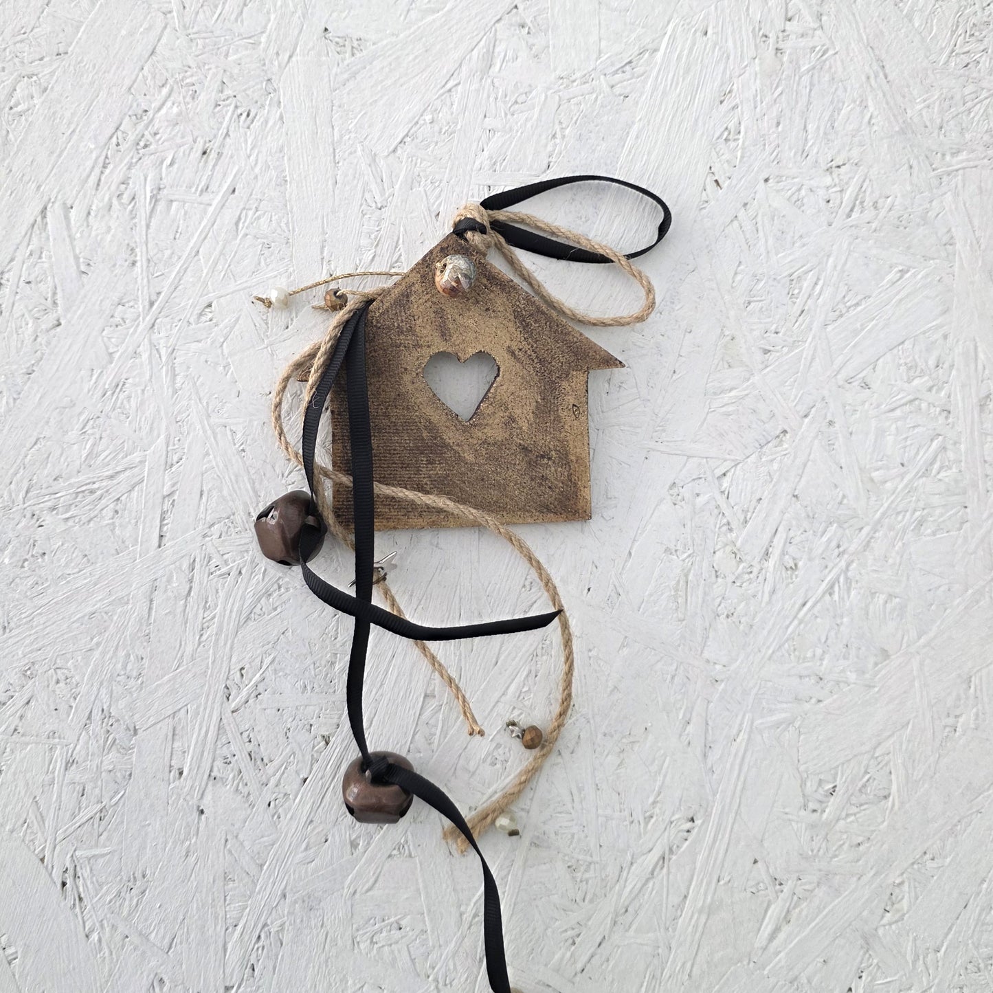 The Lucky charm House is a rustic wooden ornament shaped like a house with a heart cutout, twine and black ribbon accents, and two dark beads—perfect as a handmade ceramic-inspired Christmas decoration against any textured white backdrop.