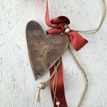 The Lucky Charm Heart features rustic metal with a twine loop, silver jingle bells, and a red ribbon bow—perfect as charming festive home decor displayed on a textured white surface.
