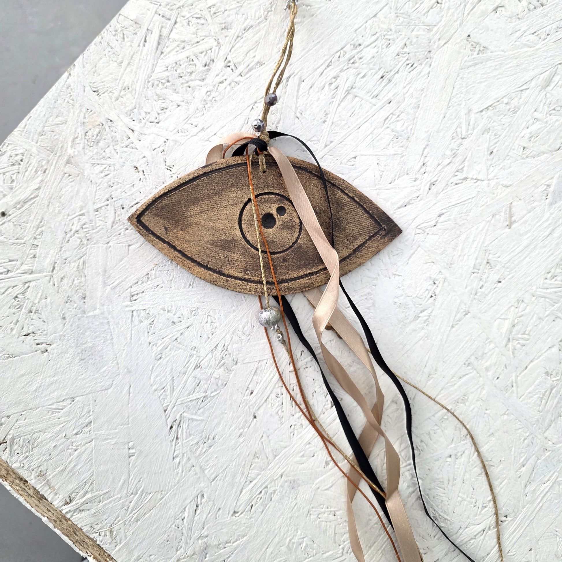 The Lucky charm Eye is a wooden eye-shaped ornament with central buttons, hanging ribbons, beads, and handmade ceramic charms inspired by Greek symbolism—perfect as a unique holiday gift.
