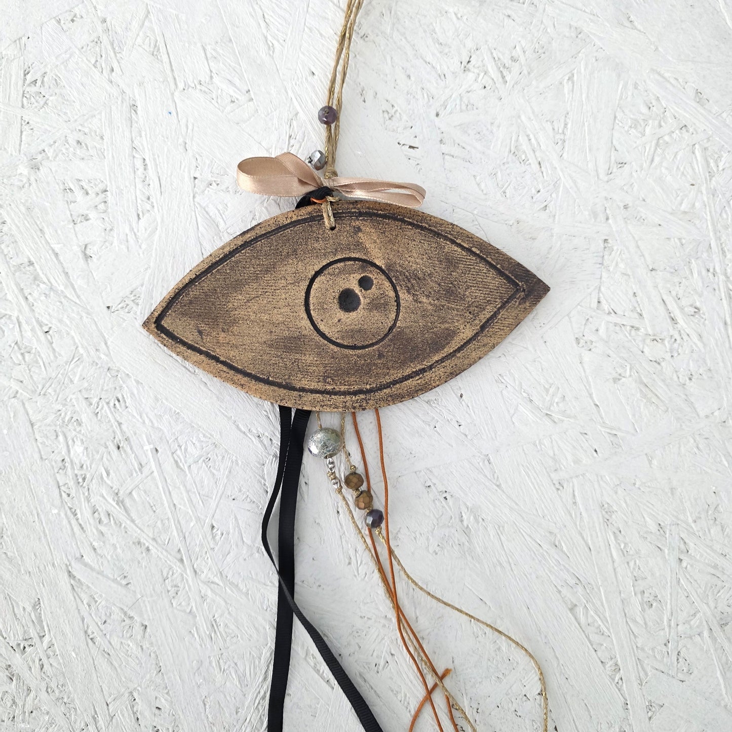 The Lucky Charm Eye, a wooden eye-shaped ornament with a circular center, hangs on a textured white wall. Featuring a peach ribbon, black and brown cords, ceramic charms, and beads, it’s ideal for holiday gifts with Greek symbolism.