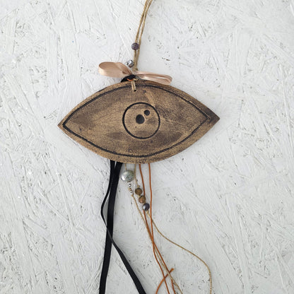 The Lucky charm Eye is a wooden, eye-shaped ornament inspired by Greek symbolism, featuring a circular center, peach ribbon, handmade ceramic charms, and beads on black and orange strings against a textured white background.