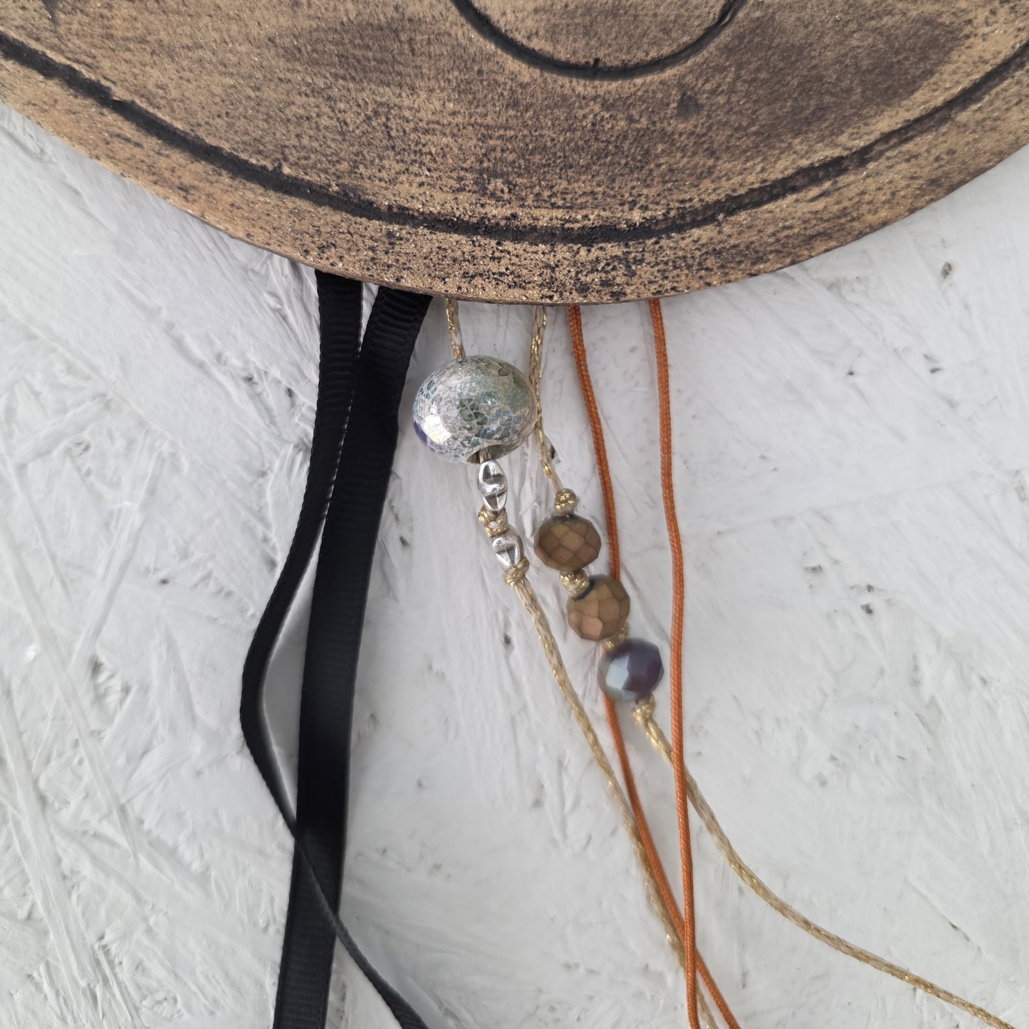 A close-up of the Lucky charm Eye featuring black, gold, and orange cords with handmade ceramic charms, hanging from a brown, round textured edge against a white textured wall.