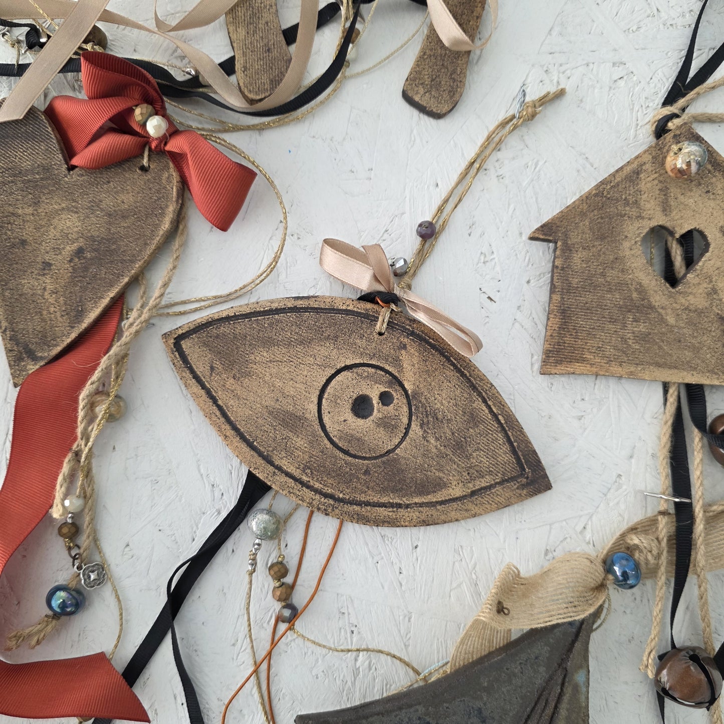 The Lucky Charm Eye, a rustic eye-shaped ornament inspired by Greek symbolism with a button center, lies on a textured white background alongside other handmade decorations like a house and heart embellished with ribbons, beads, and string.