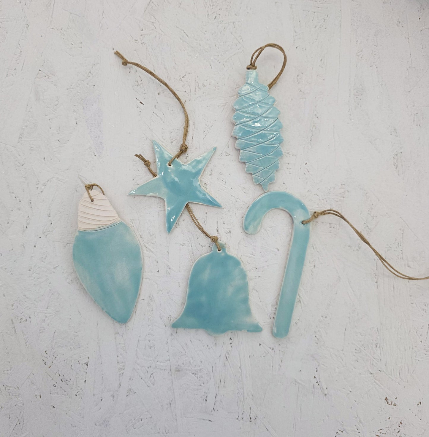Five handcrafted ceramic ornaments from Magma Lab’s Box 9—pinecone, star, seashell, bell, and candy cane—hang by brown twine on a textured white surface, making them perfect for Christmas gifting.