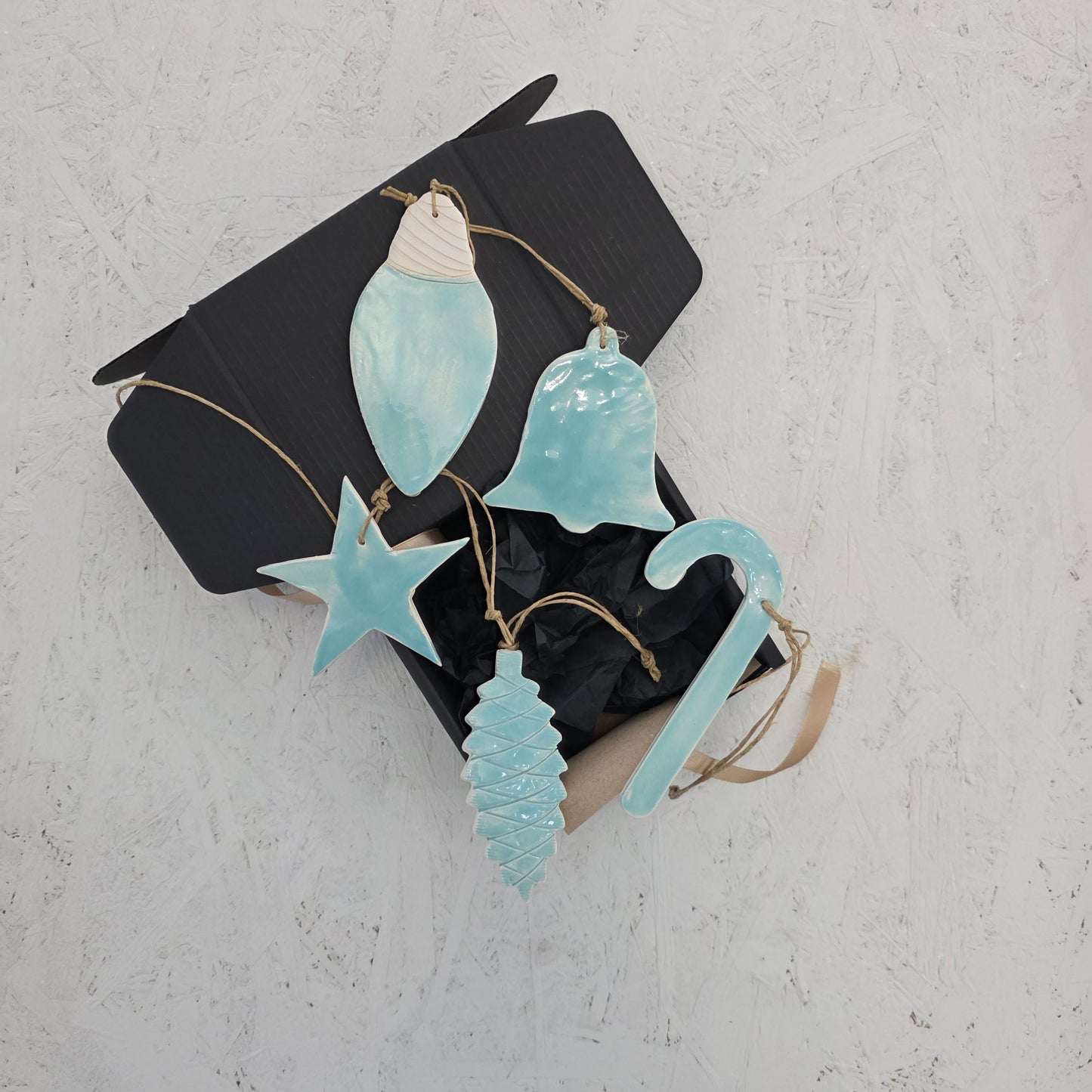 The Magma Lab Box 9 luxury gift box lies open on a white textured surface, displaying five handcrafted light blue ceramic ornaments—star, bell, Christmas tree, candy cane, and teardrop—each hanging on brown string. Perfect for Christmas gifting.