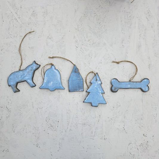 Five blue ceramic ornaments from Magma Lab’s Box 8, including a wolf, bell, house, tree, and dog bone shape, are arranged in a row on a light textured surface. These limited edition pieces feature string hangers for display.