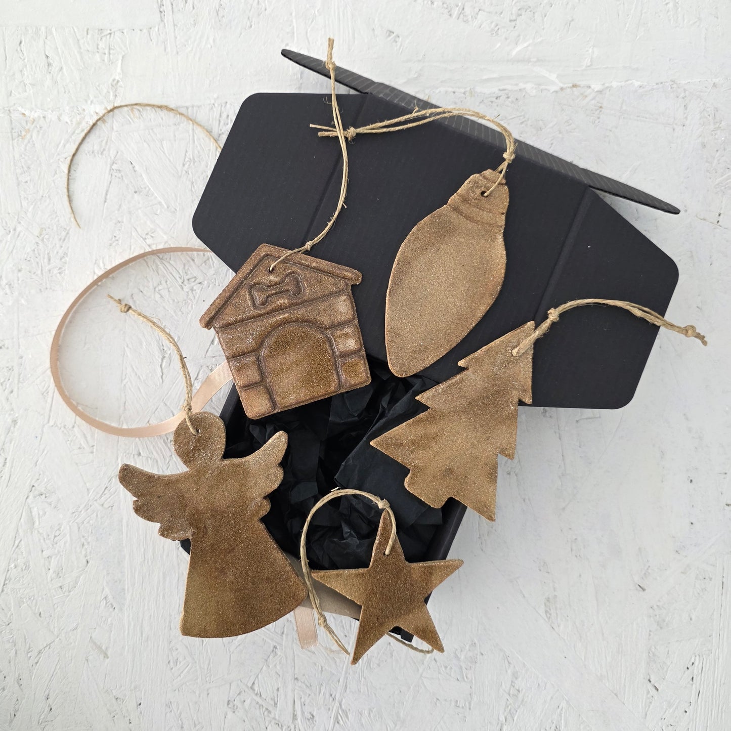 Box 10 by Magma Lab features gold handcrafted ceramic ornaments—angel, star, tree, light bulb, and doghouse—arranged with shredded black paper in a luxury gift box on a white textured surface, ideal for Christmas gifting.
