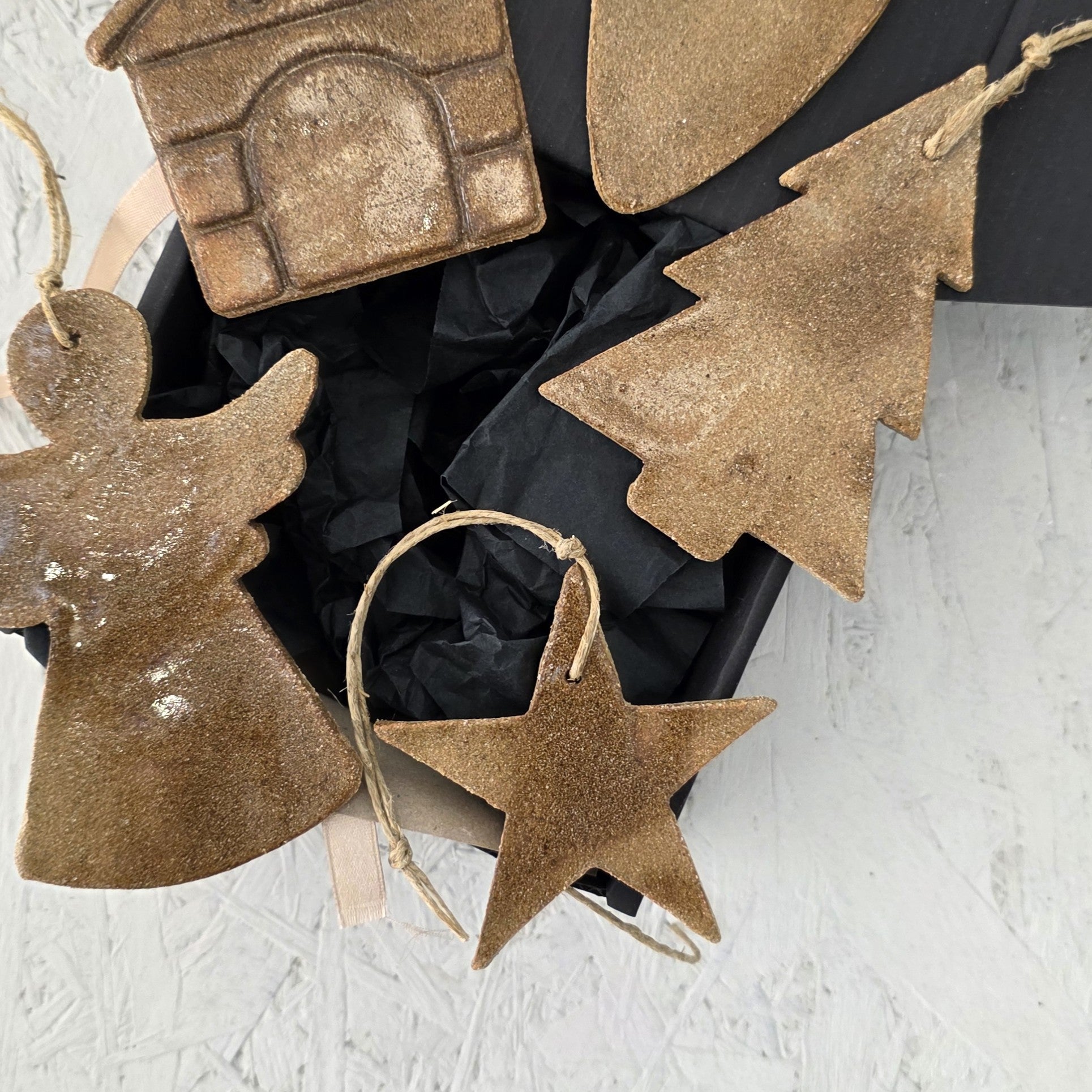 Box 10 by Magma Lab features brown, rustic handcrafted ceramic ornaments—angel, star, tree, and house—arranged on black tissue in a luxury gift box, perfect for thoughtful Christmas gifting.