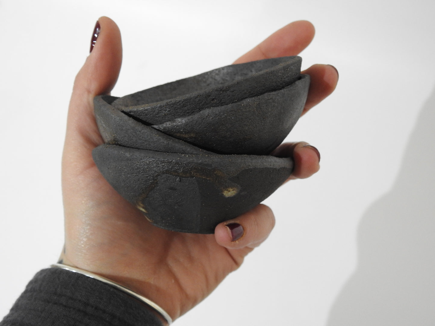 A hand holds three SOIL SMALL DIP BOWLs—small, black, roughly textured ceramics stacked together. The person wears a silver bracelet and dark nail polish. Greek craftsmanship shines against a plain white background.