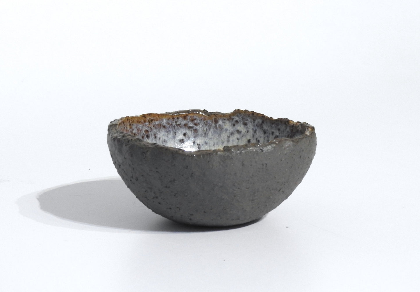 The STONE DIP BOWL is a small, handmade bowl inspired by Greek pottery, with a rough texture, uneven rim, dark gray exterior, and speckled glossy interior, shown against a plain white background.