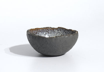 The STONE DIP BOWL is a small, handmade bowl inspired by Greek pottery, with a rough texture, uneven rim, dark gray exterior, and speckled glossy interior, shown against a plain white background.