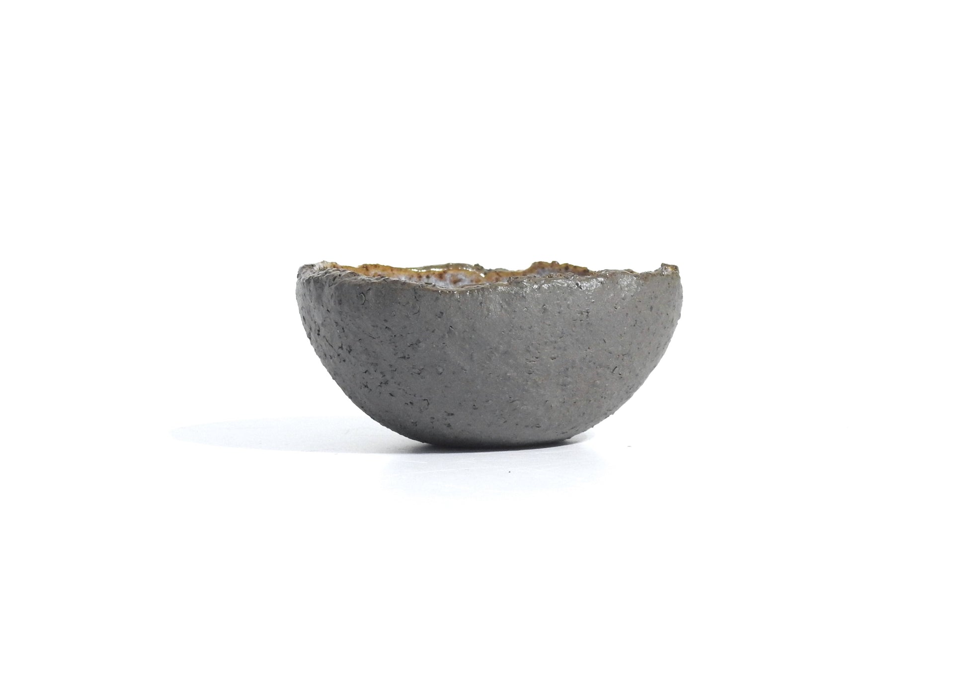 The STONE DIP BOWL is a small, gray, handmade bowl with a rough texture, uneven edges, and a minimalist rustic design inspired by Greek pottery. It is displayed on a plain white background.