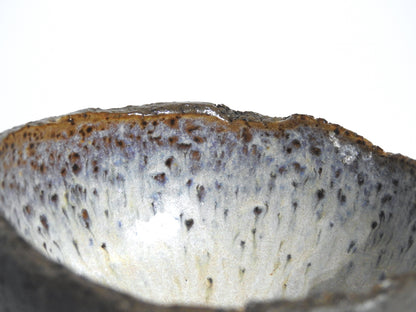 Close-up of the STONE DIP BOWL, a handmade piece with a rough, uneven rim. Its glaze blends brown, gray, and blue tones with speckled and streaked patterns, inspired by Greek pottery. Shown against a plain white background.