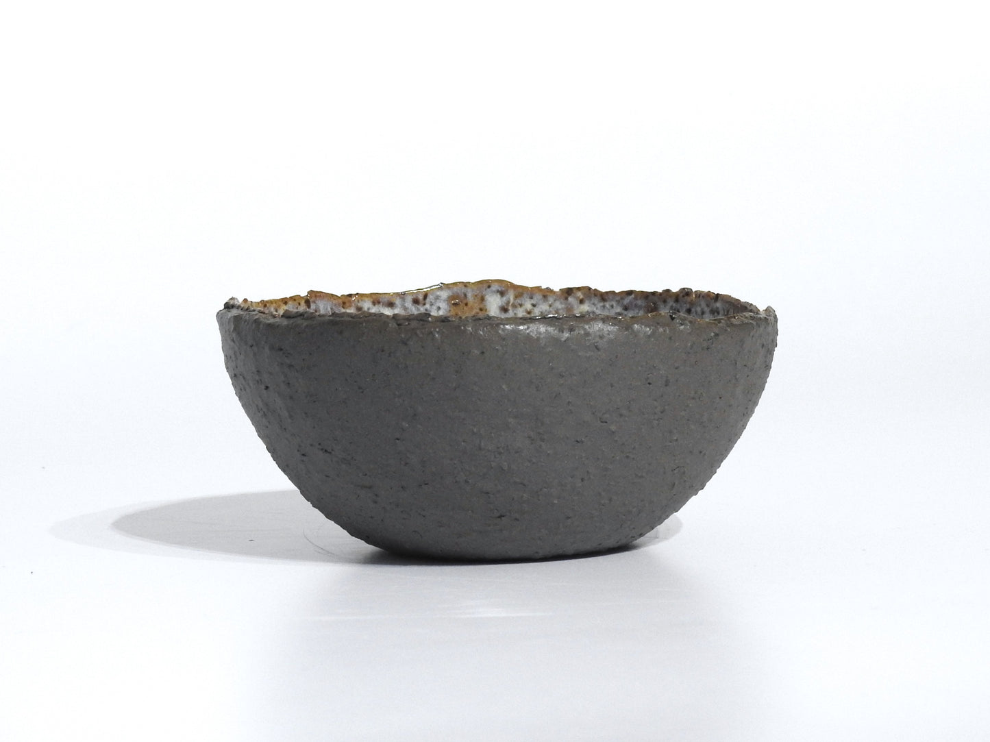 The STONE STARTER BOWL is a handmade, round grey stoneware bowl with a rough, dark exterior and slightly uneven rim, inspired by Greek pottery, shown resting on a white surface with a faint shadow.