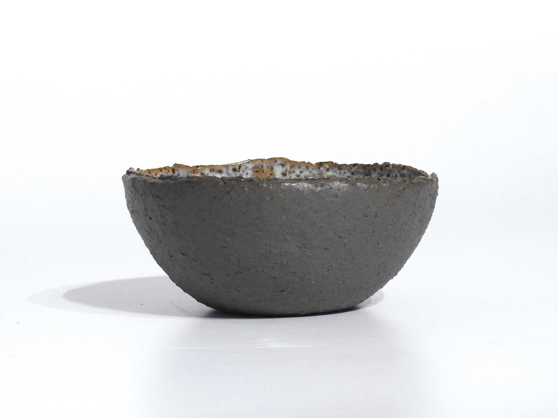 The STONE STARTER BOWL is a handmade, round grey stoneware bowl with a rough, dark exterior and slightly uneven rim, inspired by Greek pottery, shown resting on a white surface with a faint shadow.