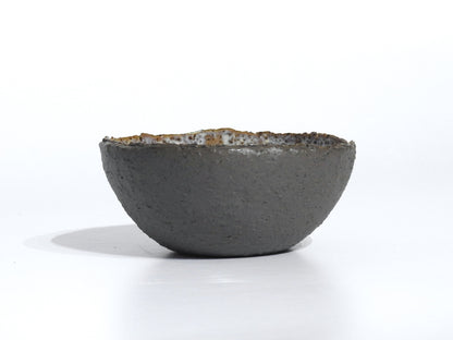 The STONE STARTER BOWL is a handmade, round grey stoneware bowl with a rough, dark exterior and slightly uneven rim, inspired by Greek pottery, shown resting on a white surface with a faint shadow.