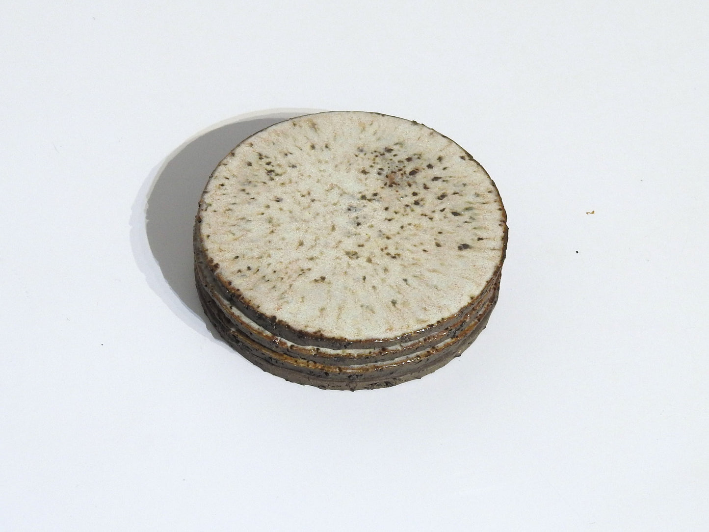 The STONE COASTERS SET OF 4 features round, flat white coasters with speckled brown spots and textured edges, resembling handmade stoneware, shown stacked on a white background.