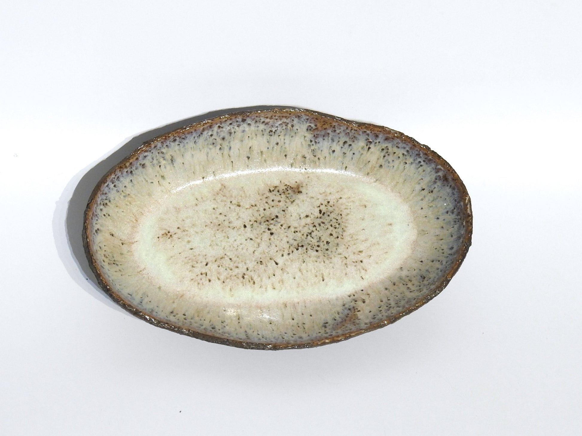 The STONE OVAL BOWL features a textured, speckled glaze in light beige and brown, measuring 15.5 x 24.5 cm. Its darker, rough rim enhances the rustic, handmade look. Shown against a plain white background.