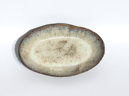 The STONE OVAL BOWL features a textured, speckled glaze in light beige and brown, measuring 15.5 x 24.5 cm. Its darker, rough rim enhances the rustic, handmade look. Shown against a plain white background.