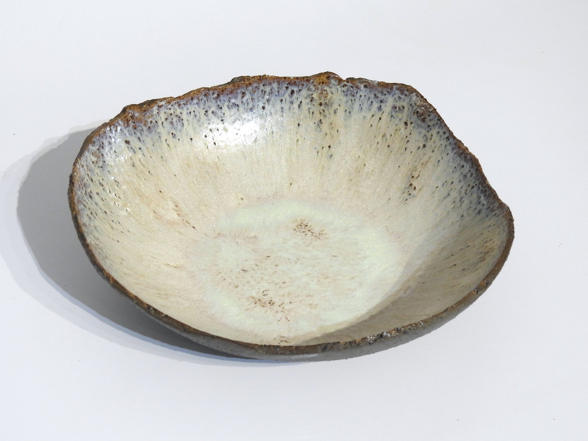 The STONE BIG SALAD BOWL is a handmade ceramic bowl with an uneven, organic rim and a speckled off-white glaze featuring brown and blue accents. Measuring 27.5 x 25.5 cm with a height of 7.5 cm, its rustic texture adds artisanal charm.