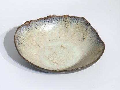 The STONE BIG SALAD BOWL is a handmade ceramic bowl with an uneven, organic rim and a speckled off-white glaze featuring brown and blue accents. Measuring 27.5 x 25.5 cm with a height of 7.5 cm, its rustic texture adds artisanal charm.