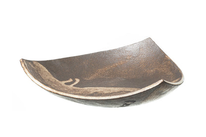 The SOIL TRIANGULAR PLATE is a handcrafted ceramic plate featuring an asymmetrical, textured brown surface and uneven curved edges, exemplifying Greek craftsmanship against a white background.