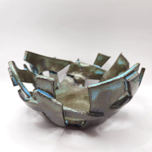 The ATLANTIS 'BRUTE' BOWL is a handmade stoneware piece with jagged, irregular edges and a fractured geometric design. Its shiny bluish-green glaze creates a unique metallic surface, shown against a plain white background.