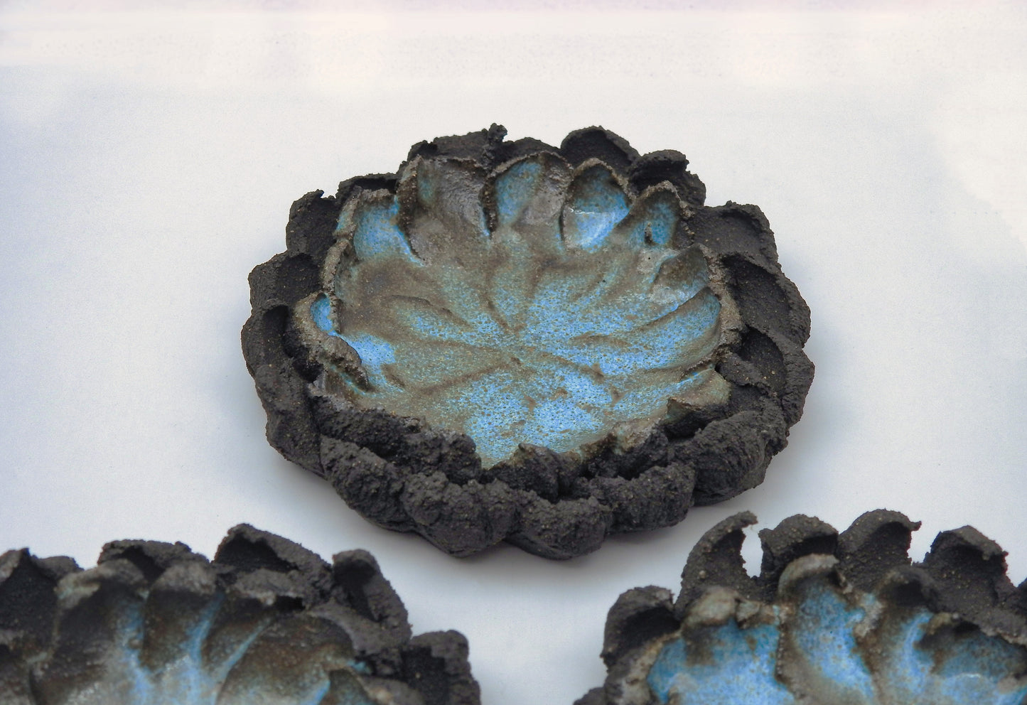 The ATLANTIS ashtray is a handmade ceramic piece with a textured dark brown, petal-shaped rim and blue speckled center. Its uneven, organic edges resemble a flower or lotus, making it an artisanal accent for any surface.