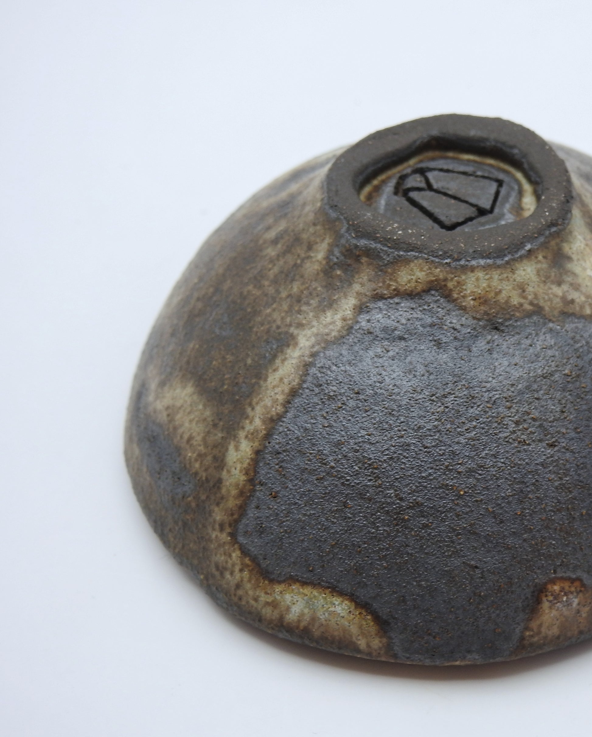 A close-up of the SOIL SMALL DIP BOWL’s rustic, textured brown and gray ceramic base highlights a maker’s mark—showcasing the beauty of handmade stoneware.