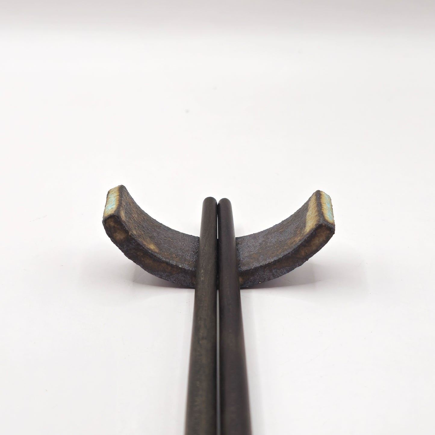 Two dark chopsticks rest on the SOIL CHOPSTICKS REST, a handmade stoneware ceramic holder with a rustic, textured finish and crescent shape, showcasing Greek craftsmanship against a plain white background.