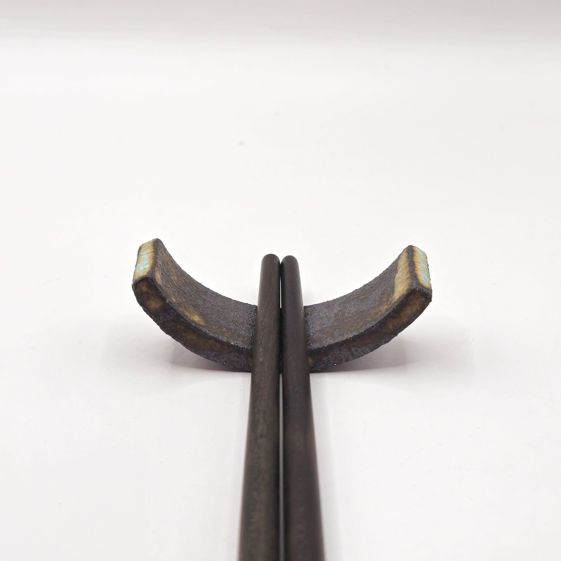 Two dark chopsticks rest on the SOIL CHOPSTICKS REST, a handmade stoneware ceramic holder with a rustic, textured finish and crescent shape, showcasing Greek craftsmanship against a plain white background.