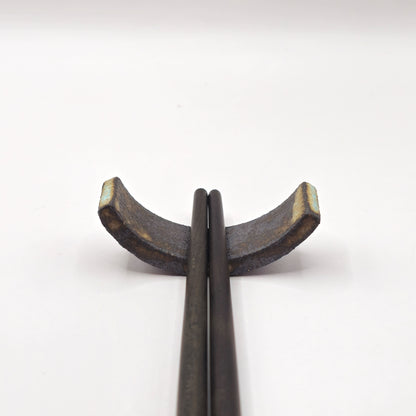 Two dark chopsticks rest on the SOIL CHOPSTICKS REST, a handmade stoneware ceramic holder with a rustic, textured finish and crescent shape, showcasing Greek craftsmanship against a plain white background.