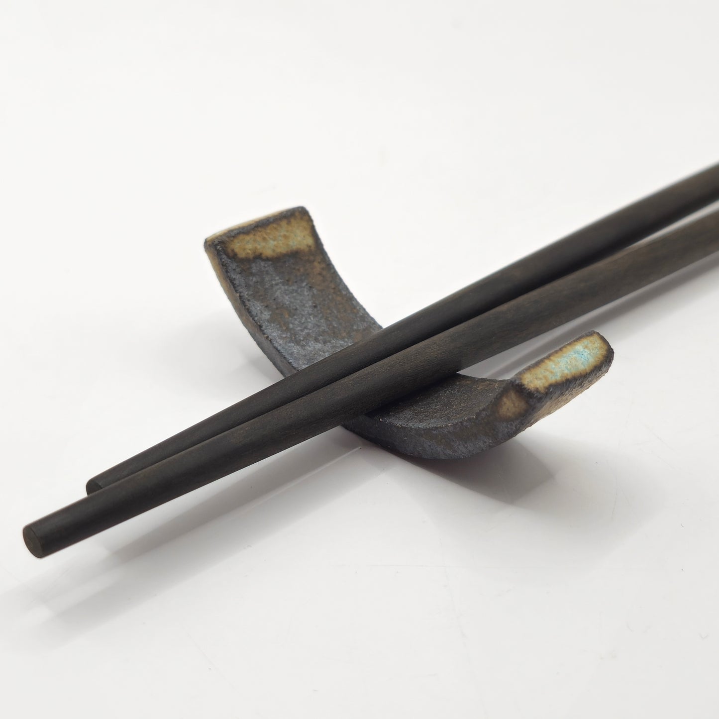 A pair of dark wooden chopsticks rests on a SOIL CHOPSTICKS REST, handmade from stoneware ceramic, set against a plain white background.