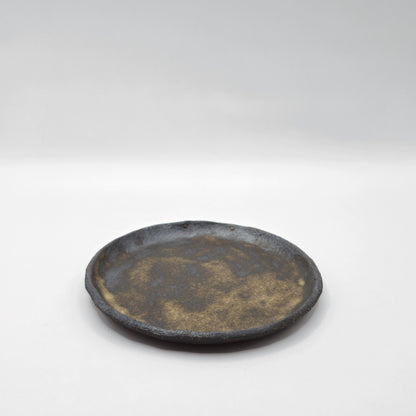The SOIL INCENSE HOLDER Set features a round, rustic ceramic plate with a textured, uneven rim and mottled gray-brown surface, handcrafted in Greek style and shown against a plain white background.