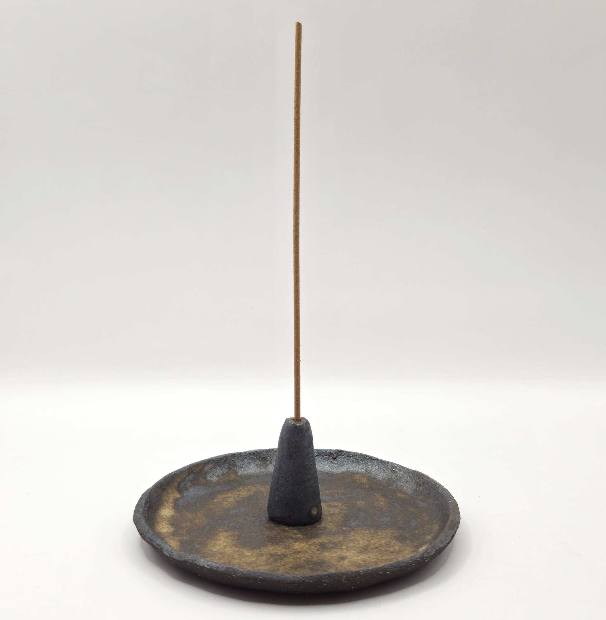 The SOIL INCENSE HOLDER Set features a slim incense stick in a handcrafted stoneware holder, displayed on a round rustic ceramic dish with earthy tones, all set against a clean white background.