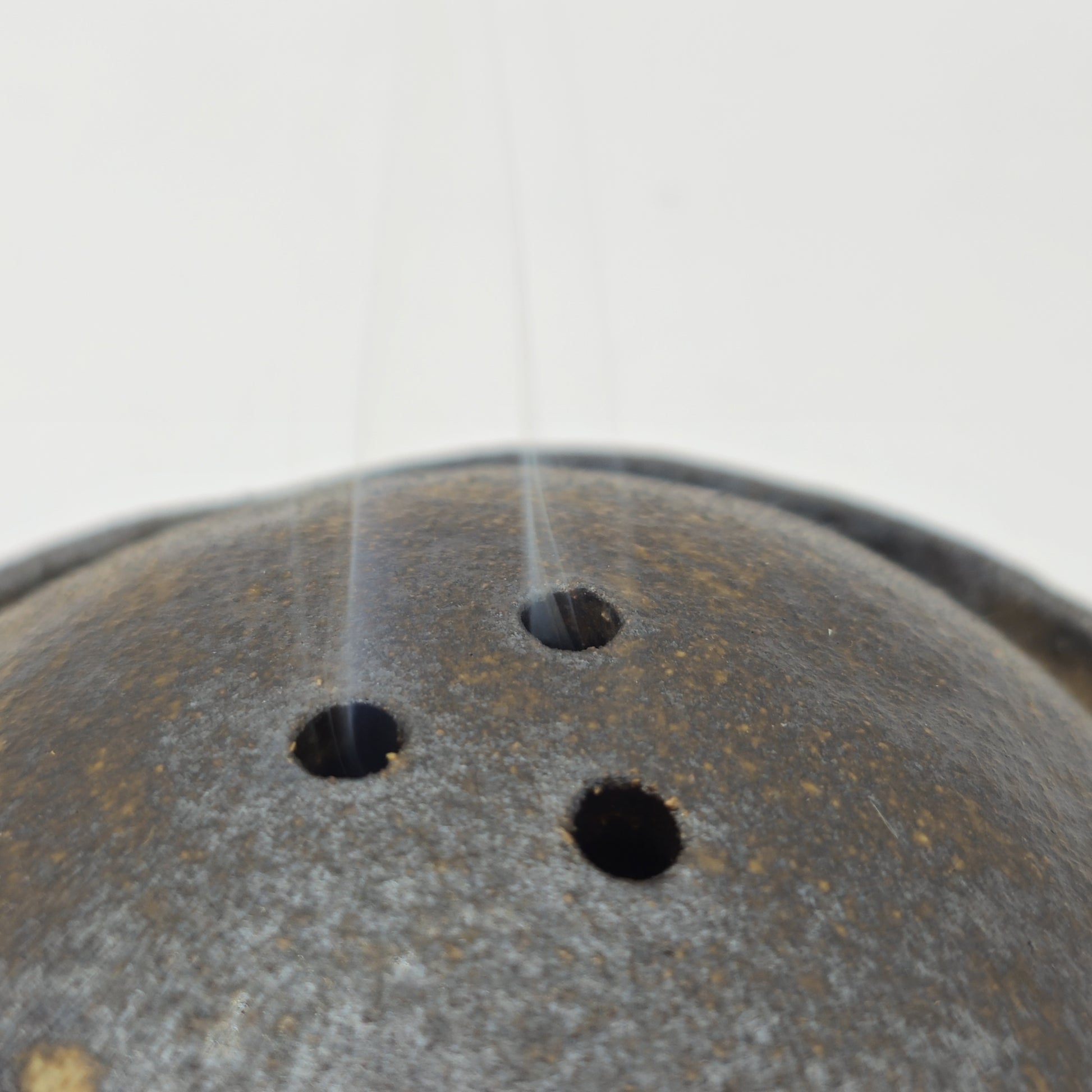 Close-up of the SOIL INCENSE HOLDER Set, a dark, rounded piece with three top holes. Thin wisps of smoke rise gracefully against a light background, highlighting its exquisite Greek craftsmanship.